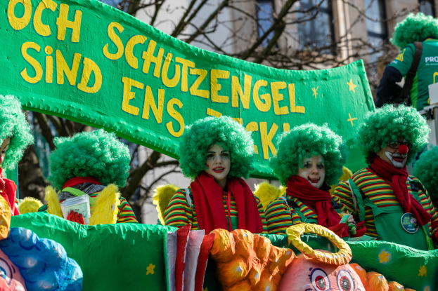 Eine Gruppe von Clowns in bunten Kostümen marschiert eine Straße entlang und hält ein Banner hoch, mit Bäumen und einem Gebäude im Hintergrund.