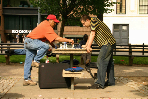 Eine Stadtansicht mit einem Tisch und Bänken, einem Schachbrett mit Figuren auf dem Tisch, Taschen auf der Bank, einem Mann mit rotem Hut und einem großen Koffer, einem anderen Mann, der auf der Bank schläft, und einem Gebäude mit Fenstern, Türen, einem Baum und Gras im Hintergrund.