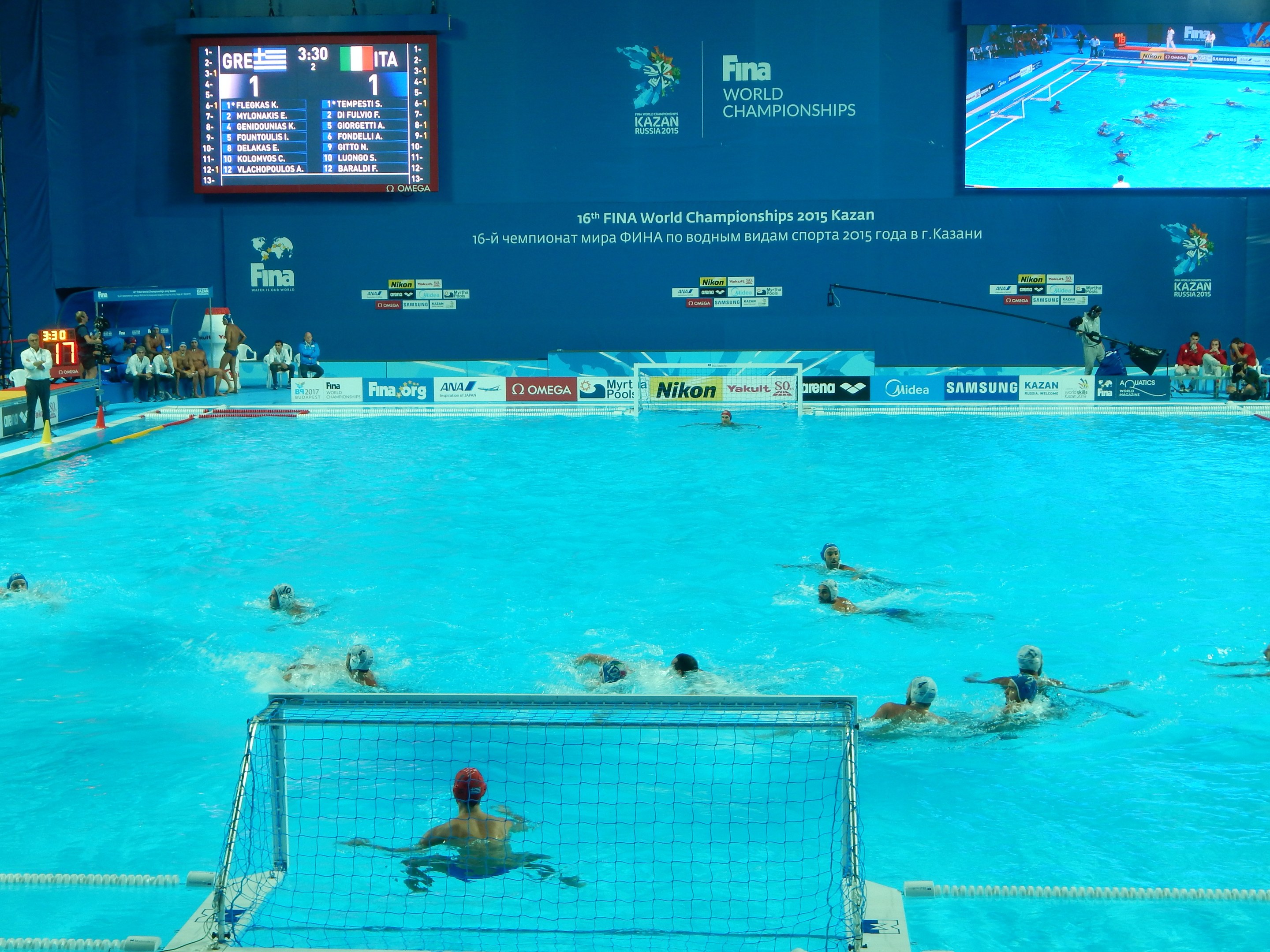 Gruppe von Menschen, die Wasserball spielen in einem Pool während der 2016 FINA Weltmeisterschaft in Kazan, mit einem Netz, das den Pool trennt und Zuschauern auf der linken Seite sitzen.