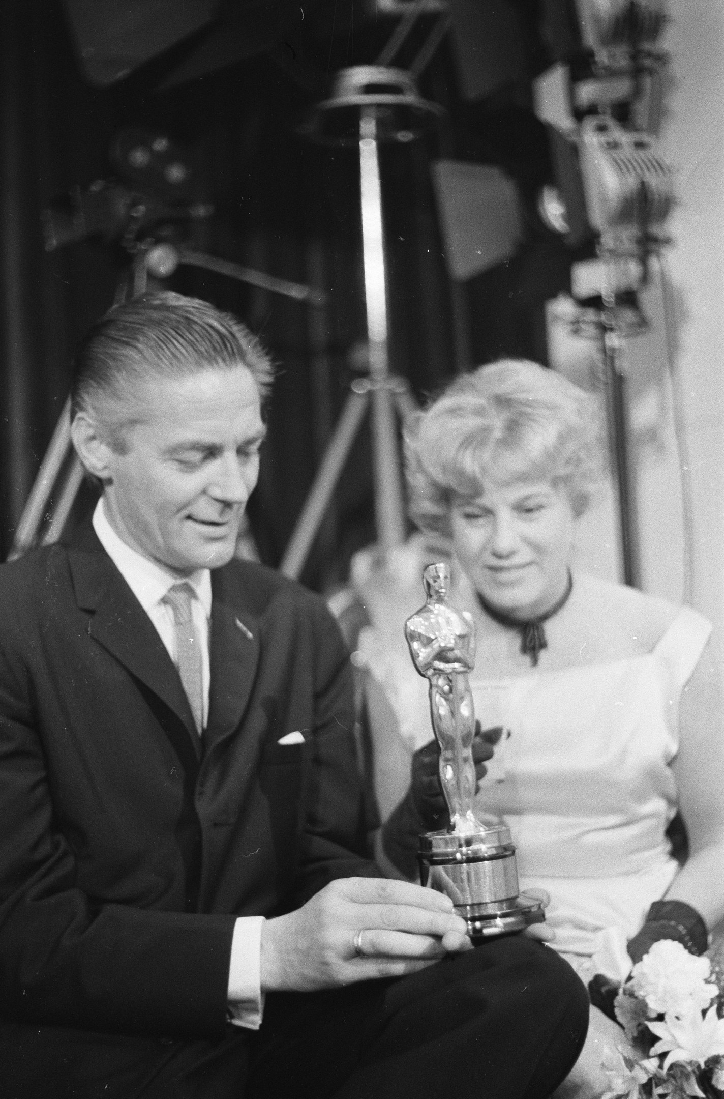 Ein Mann in einem schwarzen Blazer, weißem Hemd und Krawatte hält eine Oscar-Statue in der Hand und sitzt neben einer Frau, die einen Blumenstrauß hält, beide in Abendgarderobe, mit abgedunkeltem Hintergrund in Schwarz-Weiß.