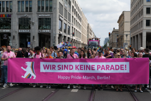 Eine Gruppe von Menschen marschiert auf einer Straße in Berlin, Deutschland, mit einem pinken "Happy Pride March"-Schild, während Gebäude, Laternenpfähle und Verkehrszeichen die Straße säumen, unter einem bewölkten Himmel.