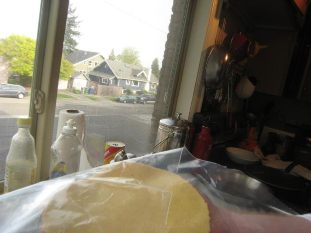 Eine Küche mit Küchenutensilien auf dem Tisch, einem Topf und Flaschen und Dosen vor einem Fenster, mit Häusern, Himmel, Bäumen, Autos und einer Straße im Hintergrund.