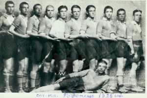 Ein Schwarz-Weiß-Foto einer Fußballmannschaft mit einem Mann in der Mitte auf dem Boden liegend und Text unten.