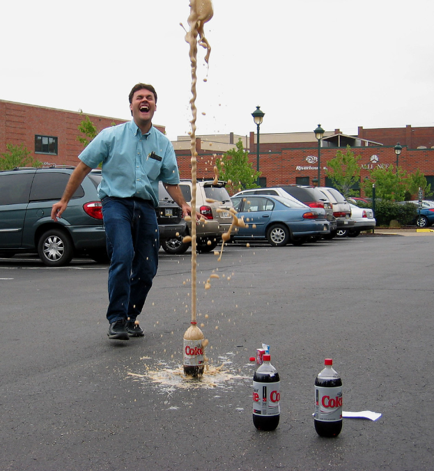 Ein Mann in einem blauen Hemd und Jeans steht lachend auf einer Straße außerhalb der Stadt, mit drei Flaschen in der Nähe, acht geparkten Autos, Bäumen, einer Straßenlaterne und einem roten Backsteingebäude im Hintergrund.