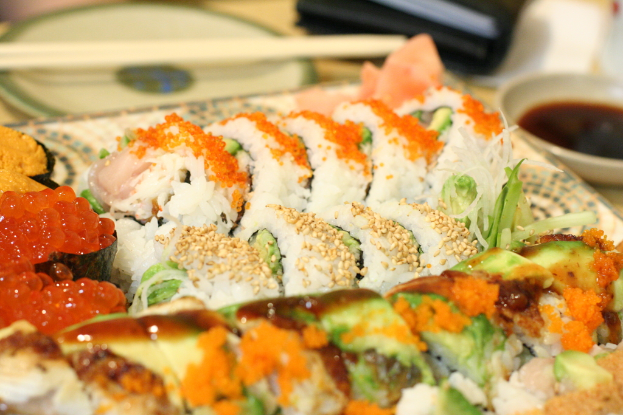 Ein Teller mit Lebensmitteln wird vor einem anderen Teller mit Stäbchen platziert, mit einer Schüssel Sauce rechts und einem schwarzen Objekt oben im Bild.