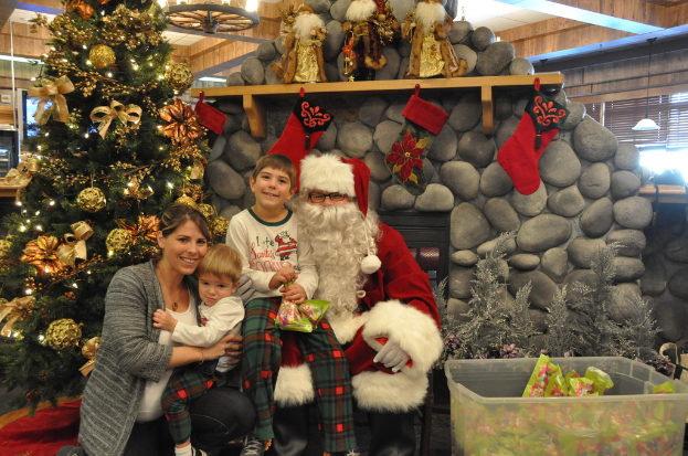 Eine Frau, Kinder und ein als Santa Claus verkleideter Mann mit Brille und Handschuhen stehen neben einem geschmückten Weihnachtsbaum, einer Steinwand, Spielzeugen und einem Korb mit Gegenständen, mit Lichtern im Hintergrund.