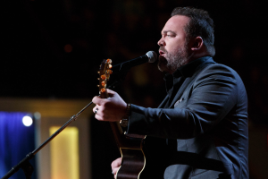 Ein Mann in einem Anzug spielt eine Gitarre und singt in ein Mikrofon auf einer schwach beleuchteten Bühne, mit Dierks Bentley im Hintergrund.