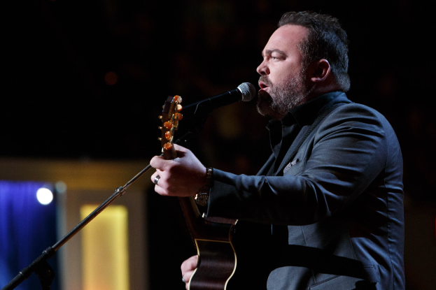 Ein Mann in einem Anzug spielt eine Gitarre und singt in ein Mikrofon auf einer schwach beleuchteten Bühne, mit Dierks Bentley im Hintergrund.