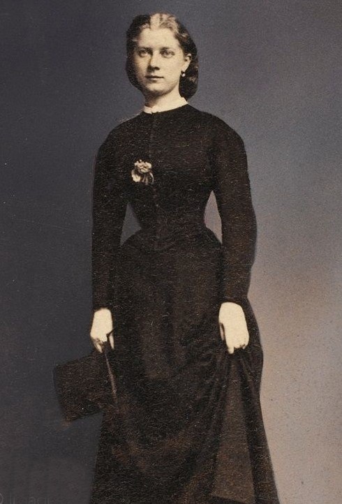 Ein Schwarz-Weiß-Foto einer älteren Frau in einem langen schwarzen Kleid mit hohem Kragen und langen Ärmeln, die eine Handtasche hält, mit einem Dutt und einem ernsten Gesichtsausdruck.