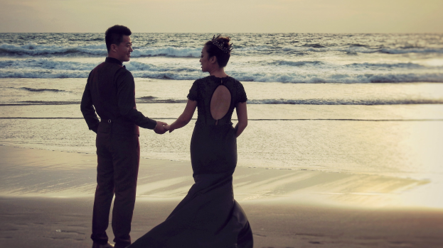 Braut und Bräutigam halten Händchen am Strand bei Sonnenuntergang, vor einem Himmel aus Orangen, Pinks und Purpurs mit tiefblauem Wasser.