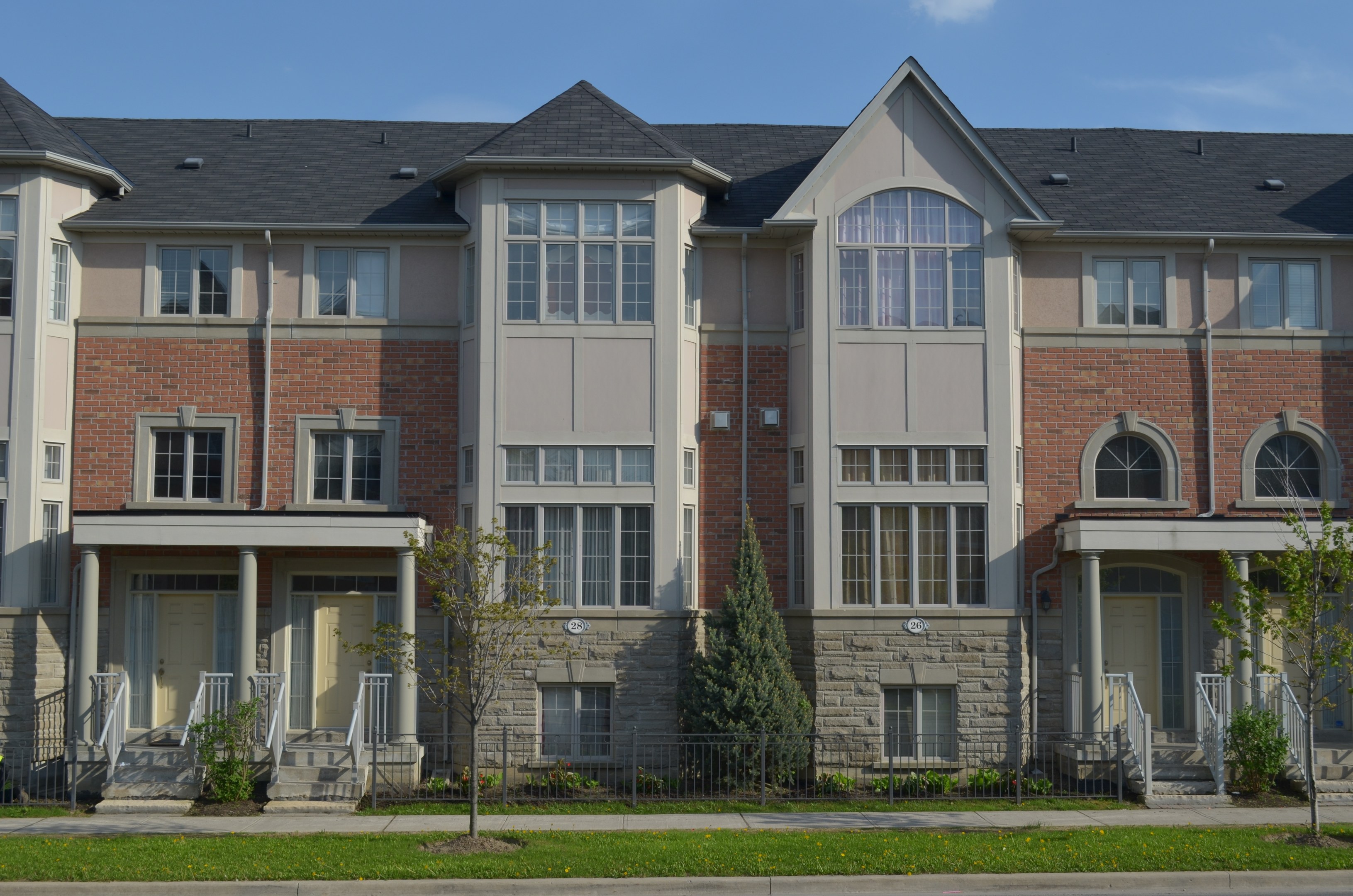 Eine Reihe von Stadthäusern in einer Wohngegend mit Fenstern, Türen, Säulen, Geländern, Treppen, Pflanzen, Bäumen, Gras, einer Straße im Vordergrund und dem Himmel im Hintergrund.