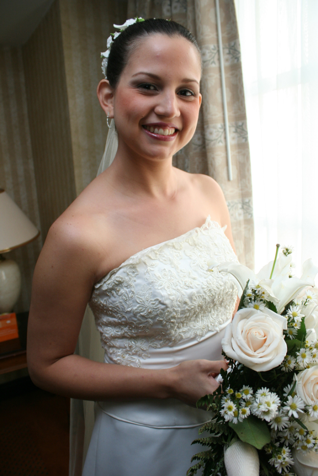 Eine Braut steht lachend und hält einen Blumenstrauß, mit einem Vorhang und einer Wand im Hintergrund und einer Lampe auf einem Tisch links.