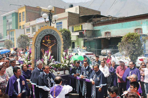 Menschen auf einer Straße mit einem Kreuz und Blumensträußen in der Mitte, Autos fahren dahinter und Gebäude, Bäume und Straßenlaternen im Hintergrund.