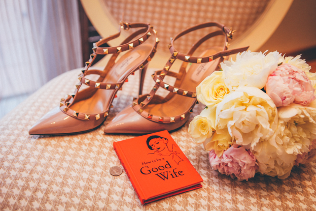 A table with a book titled "How to be a Good Wife," a bouquet of flowers, and a pair of Valentino shoes, set against a curtain backdrop.