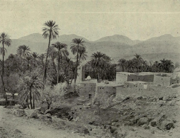 Old black and white photo of a desert village with houses, palm trees, vegetation, rocks, and mountains in the background, with text at the bottom.
