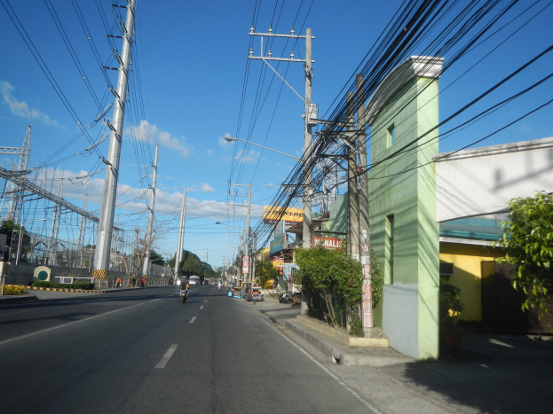 Stadtstra├če mit Stromleitungen, Fahrzeugen, Geb├Ąuden, B├Ąumen und Laternen.