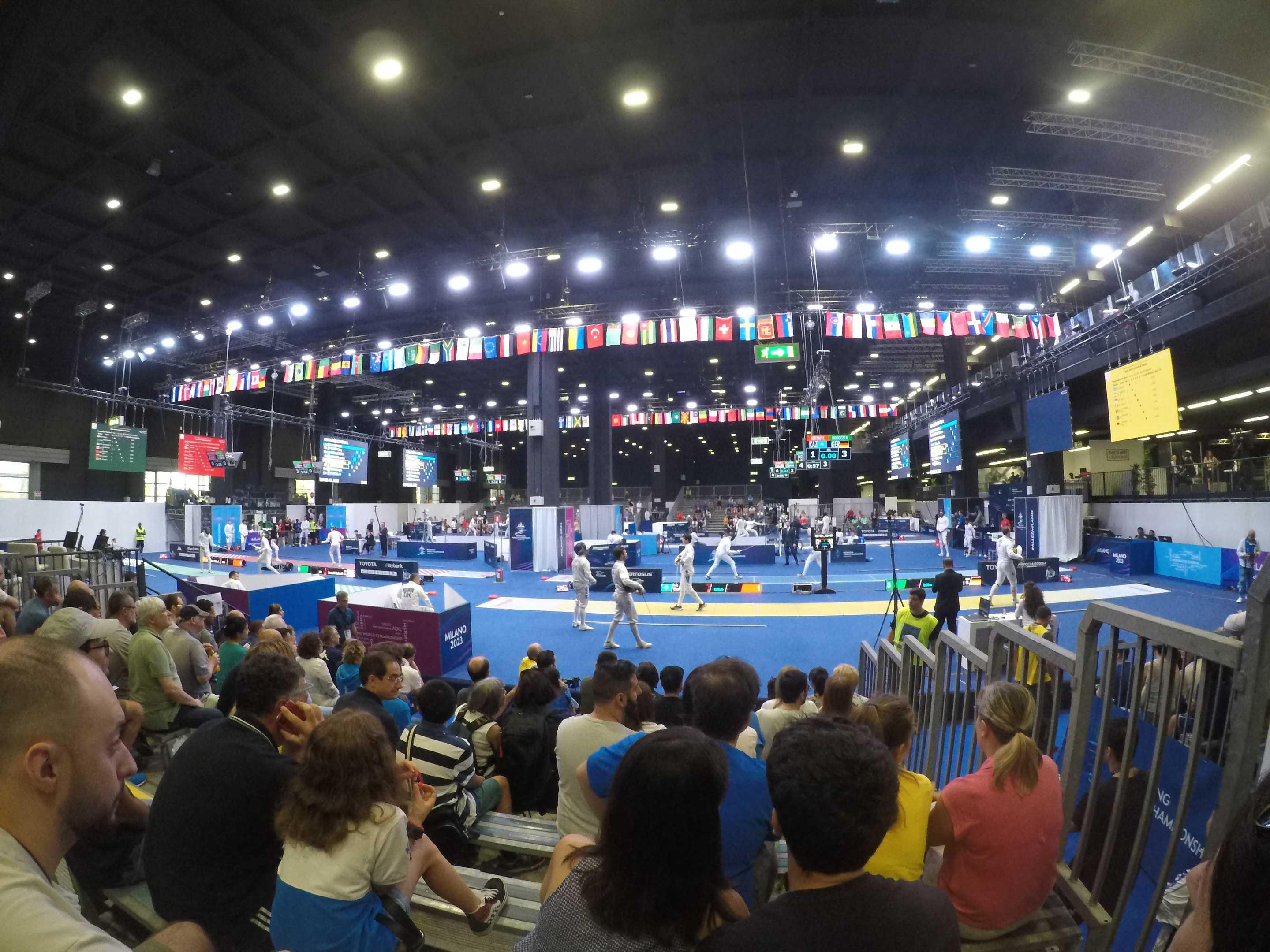 A large crowd sits facing a stage with a railing on the right and overhead lights, while a few people stand on the stage during the 2018 World Championships in Munich, Germany.
