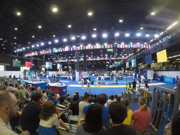 A large crowd sits facing a stage with a railing on the right and overhead lights, while a few people stand on the stage during the 2018 World Championships in Munich, Germany.