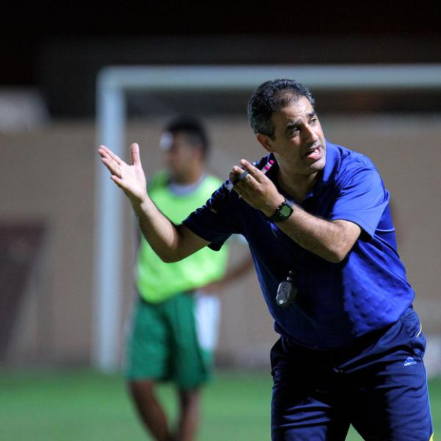 Ein Mann in einem blauen T-Shirt und Hose steht auf einem Fußballfeld und hält ein Mikrofon in der Hand, während er auf seine Spieler zeigt. Im Hintergrund ist eine Person in einem grünen Kleid, ein Torpfosten und eine Wand zu sehen.