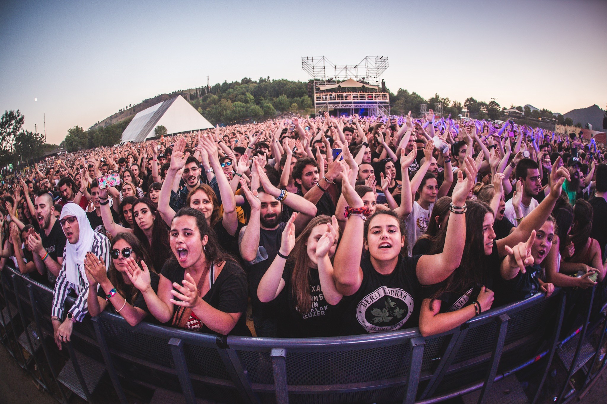 A large crowd at a music festival with raised hands in front of a railing, trees, buildings, and a clear blue sky in the background.