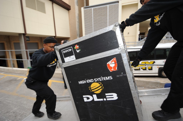 Gebäude mit einem Bus, zwei Männer in schwarzen Gewändern und Mützen mit einer Kiste.