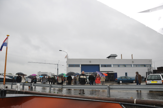 Eine Gruppe von Menschen, die draußen stehen, einige halten Schirme, mit einer Fahne an einem Stab, zwei Autos, ein Gebäude mit Fenstern, ein Turmkran, eine Straßenlaterne und eine Barrikade.