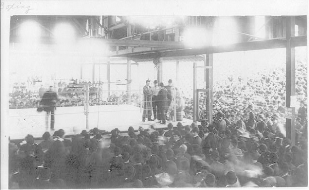 Schwarzes und weißes Foto einer Menge in einer Stadion, die einen Boxkampf beobachten, mit wenigen Menschen auf der Bühne und anderen im Publikum.