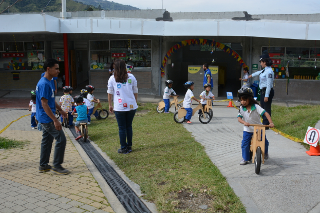 Kinder mit Helmen fahren Fahrräder auf einer Rasenfläche, mit Menschen in der Nähe und einem Schild und einem Gebäude im Hintergrund.