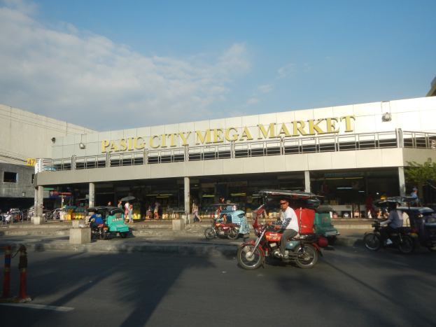 Eine belebte Straßenszene am Pasig City Mega Market mit Fahrzeugen und Fußgängern, ein Gebäude mit Text im Hintergrund und ein bewölkter Himmel.