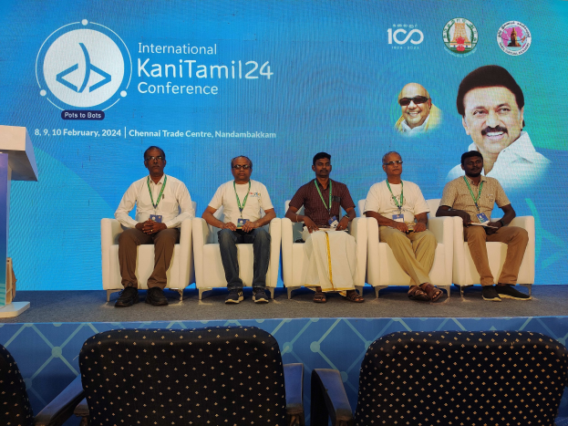 Gruppe von Menschen, die auf Stühlen vor einem Podium mit Mikrofon sitzen, mit einem Banner im Hintergrund, das Text und Bilder zeigt.