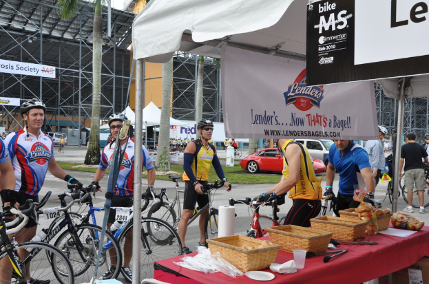 Eine Straßenszene mit Menschen, die in der Nähe von Fahrrädern stehen, ein Geschäft mit Gegenständen auf einem Tisch, Bäume, Zelte, an eine Hütte angehängte Banner, Fahrzeuge und Gras.