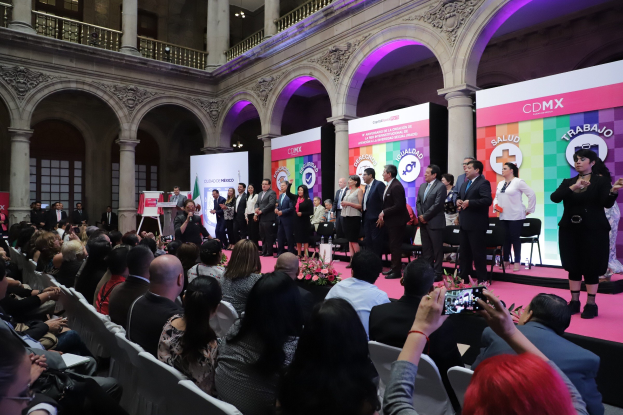 A group of people standing on a stage in front of a seated audience, with banners, pillars, railings, and lights in the background, some holding cameras.