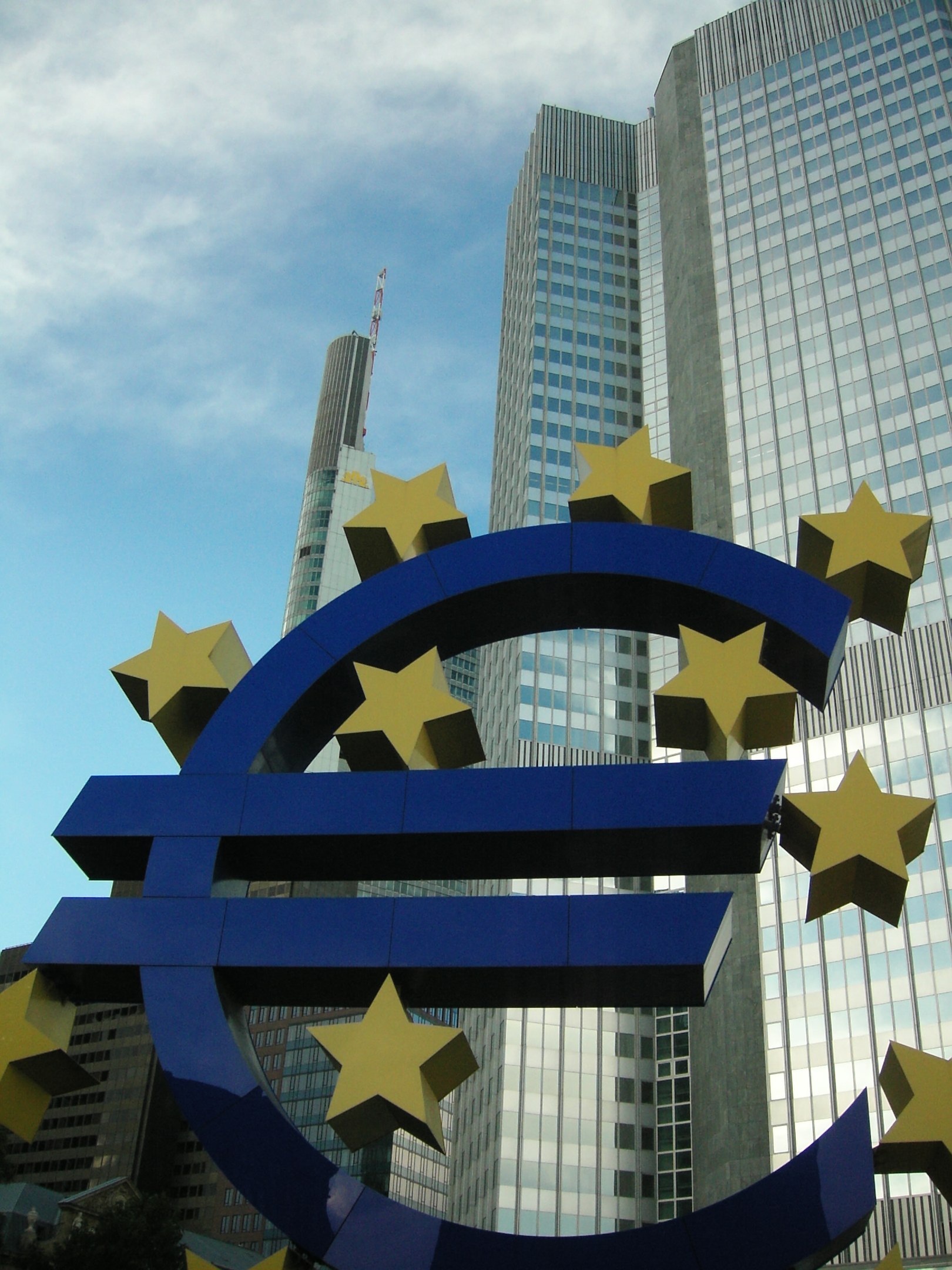 Euro-Zeichen vor dem Hauptquartier der Europäischen Zentralbank in Frankfurt, Deutschland, mit Bäumen und einem bewölkten Himmel.