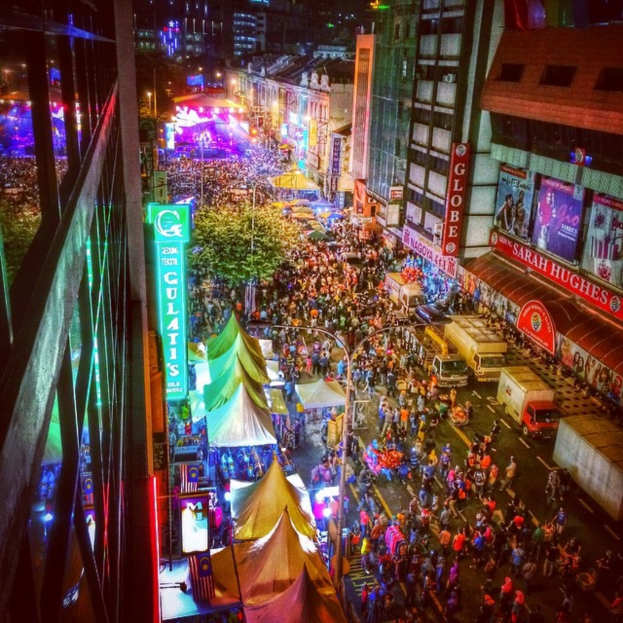 Eine belebte Jahrmarktszene mit einer groÃen Menge auf der StraÃe, umgeben von Zelten, GeschÃ¤ften, GebÃ¤uden und Bannern, mit zahlreichen Lichtern im Hintergrund.