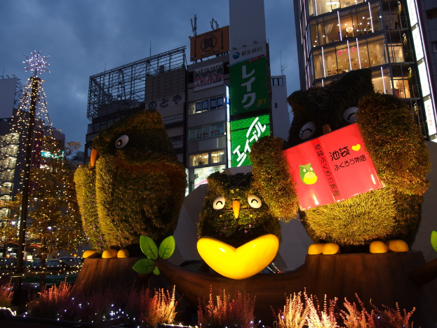 Ein Weihnachtsbaum mit Schmuck, eine Glaswand, Lichter, Säulen, ein Plakat und drei Personen vor einem Gebäude.