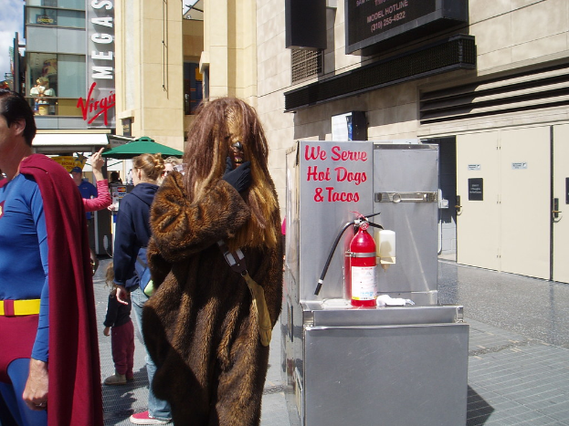 Eine Gruppe von Menschen, einige in Kostümen, versammelte sich vor Gebäuden mit sichtbarer Beschriftung, einer Box mit Schrift und einem Feuerlöscher.