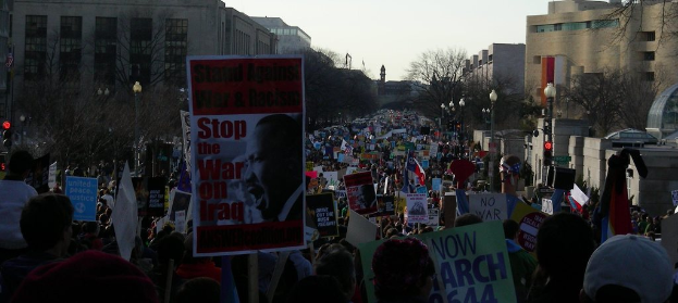 Menschen mit Plakaten auf einer Straße protestierend, mit Gebäuden, Bäumen, Laternenmasten und Himmel im Hintergrund.