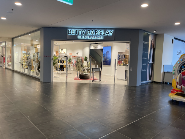 Interior of a Betty Barclay store in a shopping mall displaying mannequins in dresses, hanging clothes, tables, plants, and a toy vehicle on the right, with a "Betty Barclay Selected Brands" sign above.