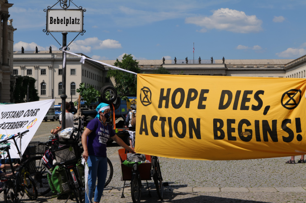 Eine Gruppe von Menschen steht vor einem Gebäude und hält ein gelbes Banner mit der Aufschrift "Hope Dies, Action Begins". In der Nähe stehen Fahrräder und ein Pfahl mit einer Tafel, unter einem klaren blauen Himmel mit Bäumen im Hintergrund.
