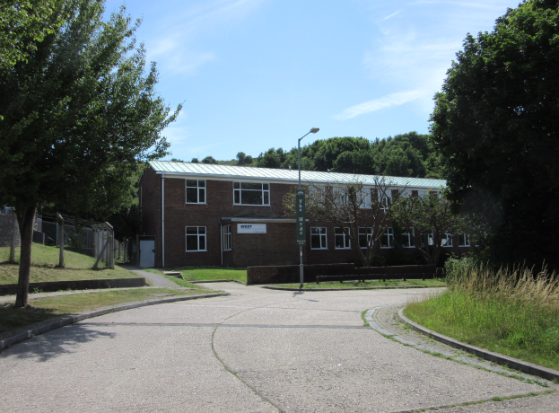 Ein Schulgebäude mit Fenstern, einer Straßenlaterne, einem Schild, Bäumen, Gras, einem Zaun und einem bewölkten Himmel, das sich in einem Park mit saftigem Grün befindet.