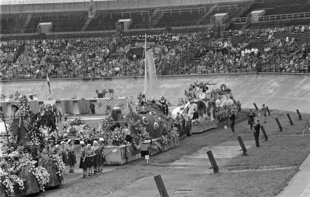 Schwarzes und weißes Foto eines Umzugs in einem Stadion mit Menschen, die stehen und sitzen, einem Brunnen in der Mitte und Blumensträußen an den Seiten der Fahrzeuge.