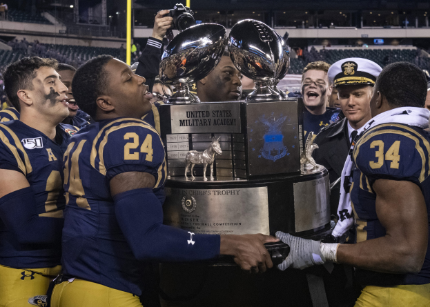 A group of men holding a trophy in a brightly lit stadium, with one man in the center photographing the celebration.