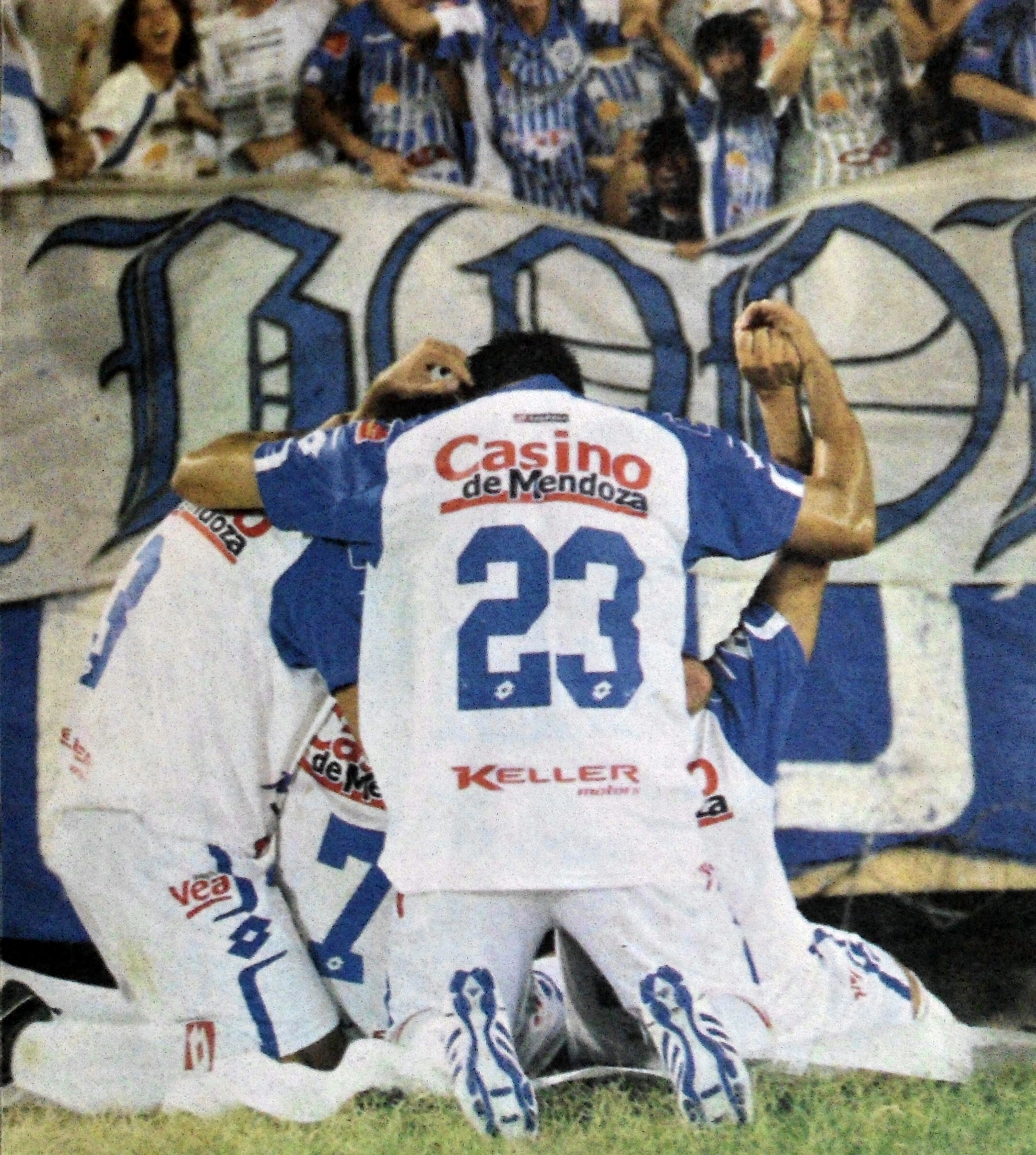 Eine Gruppe von Fussballspielern feiert ein Tor auf dem Platz vor einer jubelnden Menge, mit einem Banner mit Text im Hintergrund.