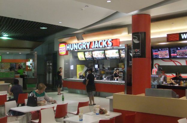 In einem Restaurant stehen Frauen an einer Essensbestellung auf einer Seite, während auf der anderen Seite Tische und Stühle mit einer Frau und einem Mann an einem der Tische angeordnet sind.