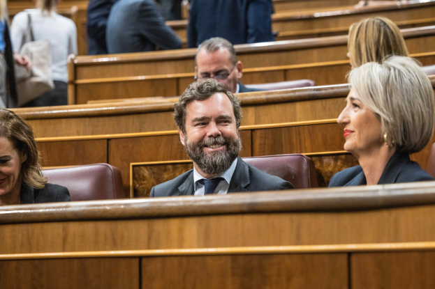 Gruppe von Menschen, die in einem Gerichtssaal sitzen, mit einem Mann in einem Anzug und zwei leger gekleideten Frauen, die nebeneinander sitzen, sowie sichtbaren Bänken im Hintergrund.