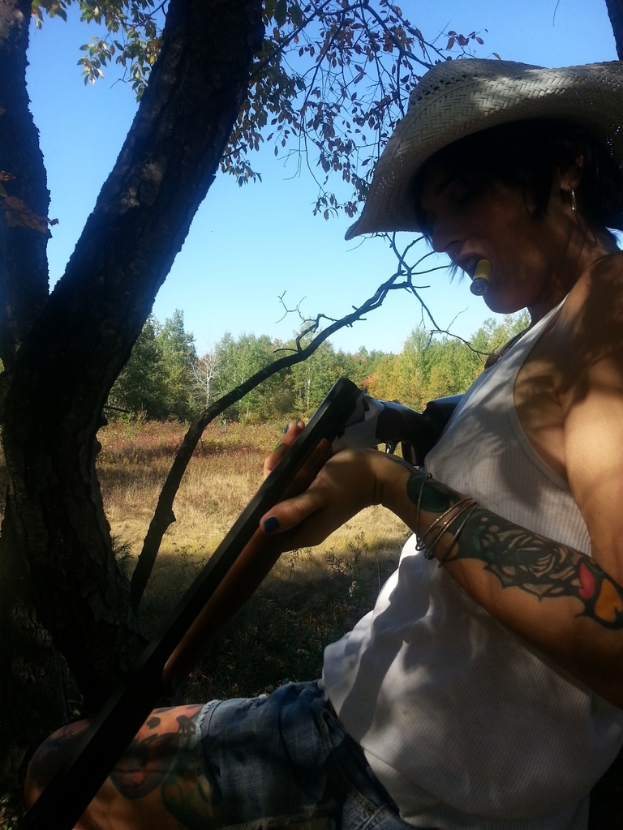 Eine Frau steht mit einer Waffe in der Hand und einer Zigarette im Mund umgeben von Bäumen, Gras und Himmel.