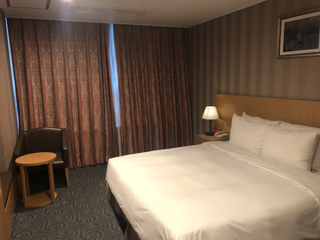 Hotelzimmer mit einem großen Bett, Stuhl, Tisch, Telefon, Lampe, Foto auf der Wand, Fenster mit Vorhängen und Deckenbeleuchtung.