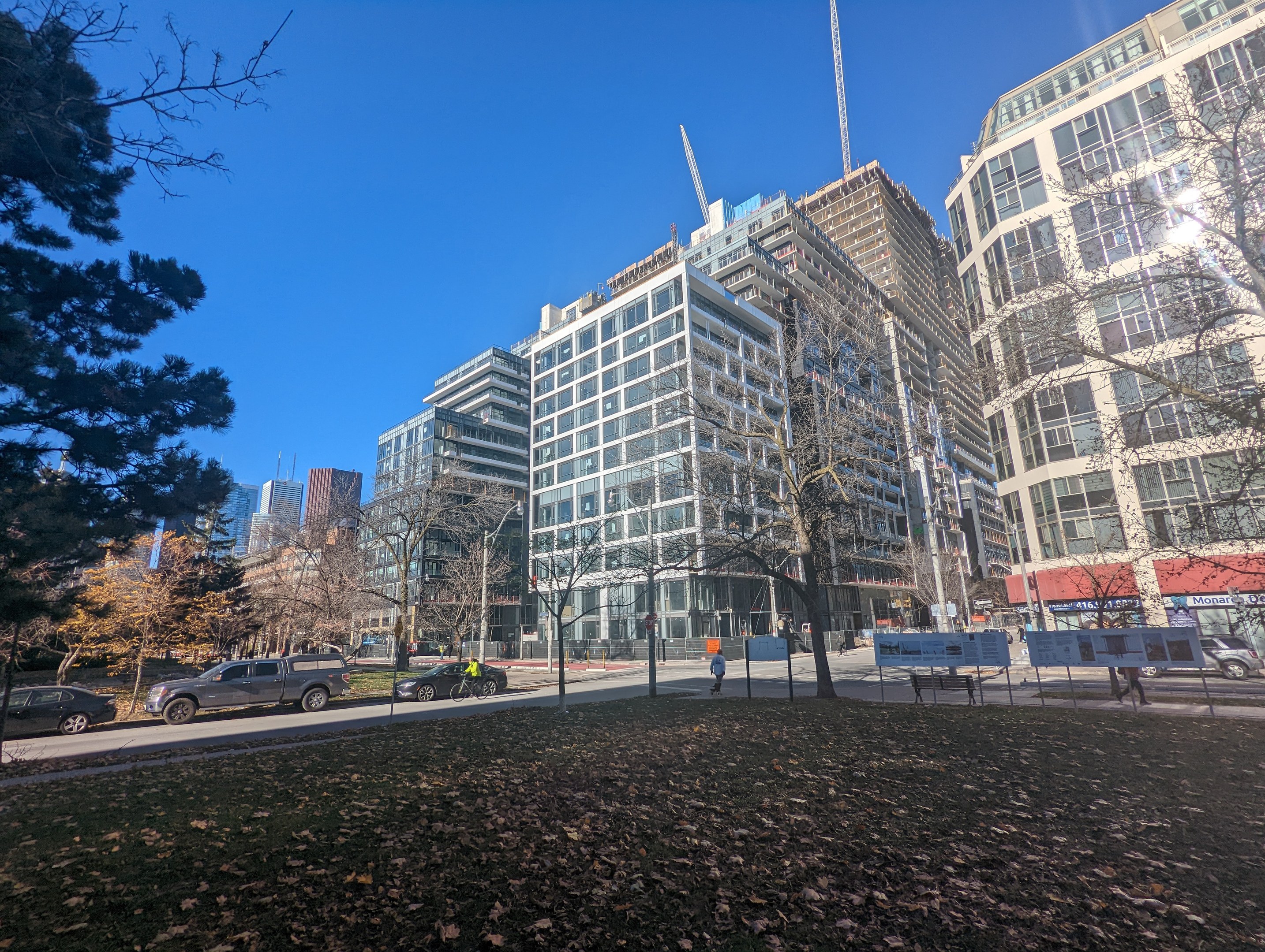 A city street in Toronto with tall buildings, moving vehicles, pedestrians on the sidewalk, trees, and scattered dry leaves on the ground under a visible sky.