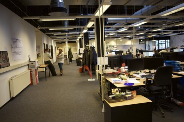 Ein geräumiges Großraumbüro mit zahlreichen Schreibtischen, Stühlen und Computern, Menschen bei der Arbeit, verschiedenen Gegenständen auf den Tischen, hängenden Kleidern, Wanddisplays und Deckenleuchten, mit Glasfenstern im Hintergrund.