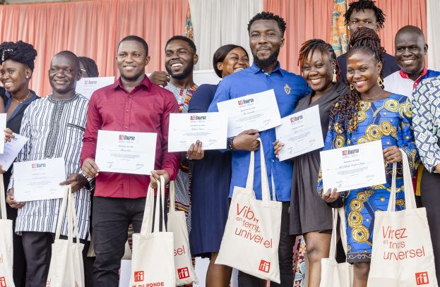 A group of people smiling and holding certificates, with bags at their feet, standing near curtains and a wall.
